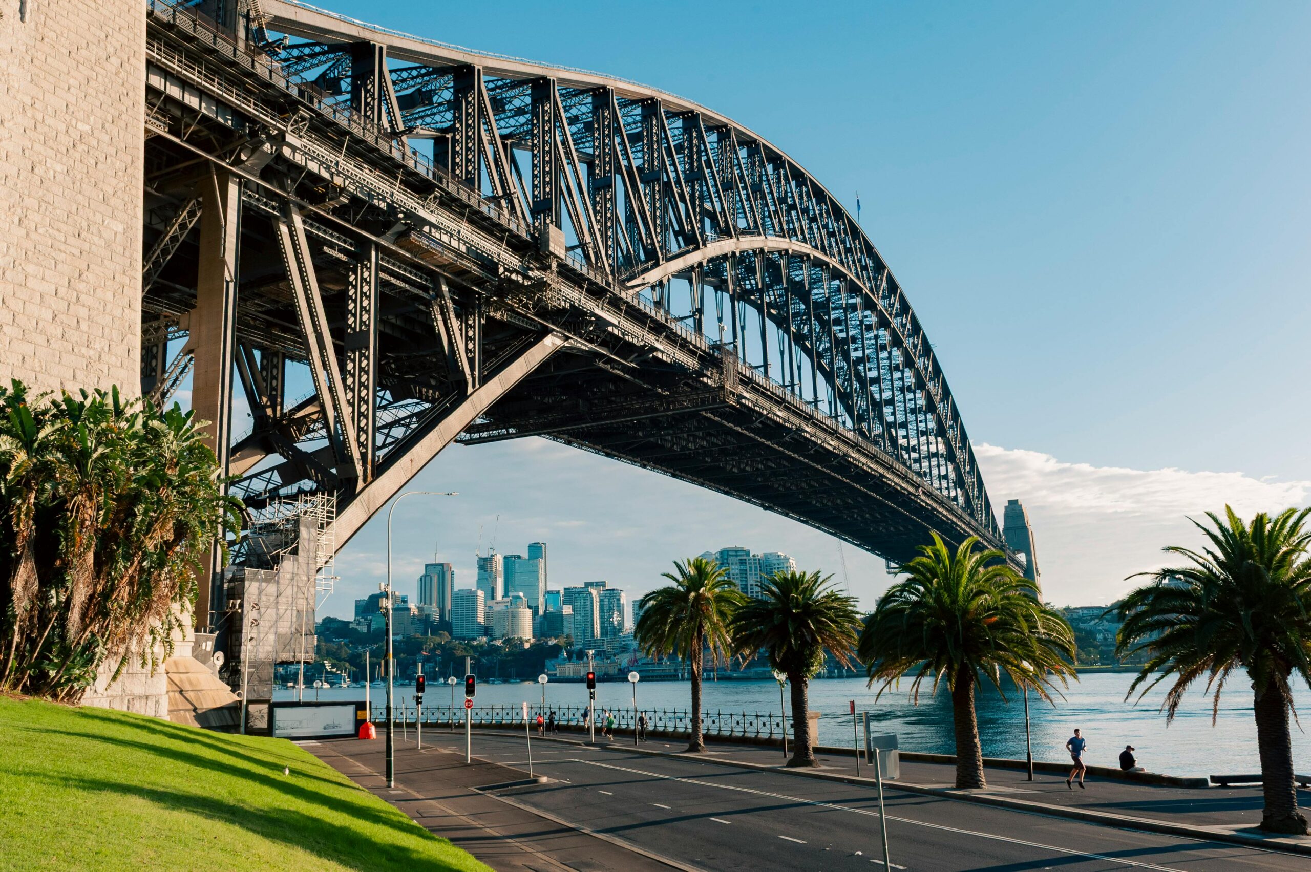 A scenic view of Sydney Harbour Bridge with clear blue sky and palm trees along a quiet road.
